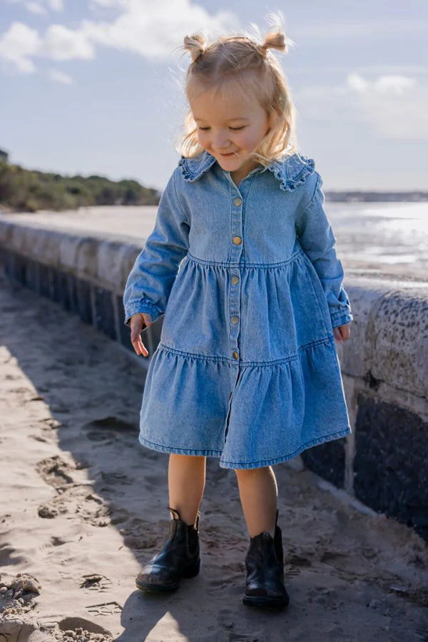 Young girl in a blue denim dress standing on a beach.