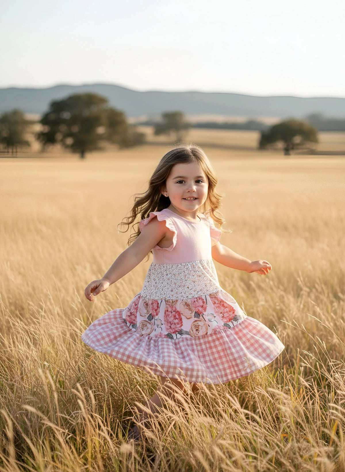 A young girl with long brown hair runs and twirls in a golden wheat field, wearing a pink sleeveless dress with a white crochet yoke and a pink gingham skirt with floral applique.