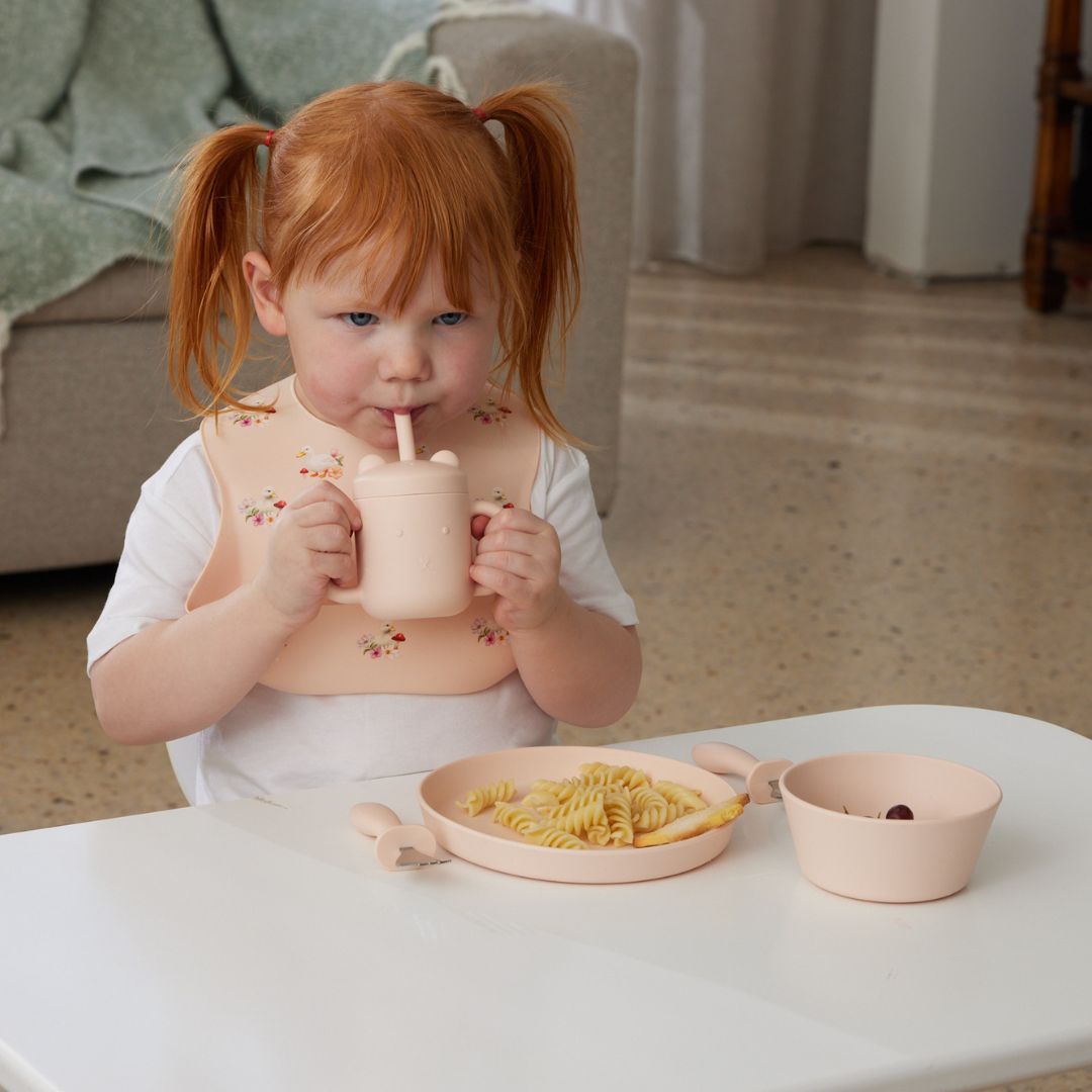 Child drinking from a cup with a straw, sitting at a table with a plate of food.