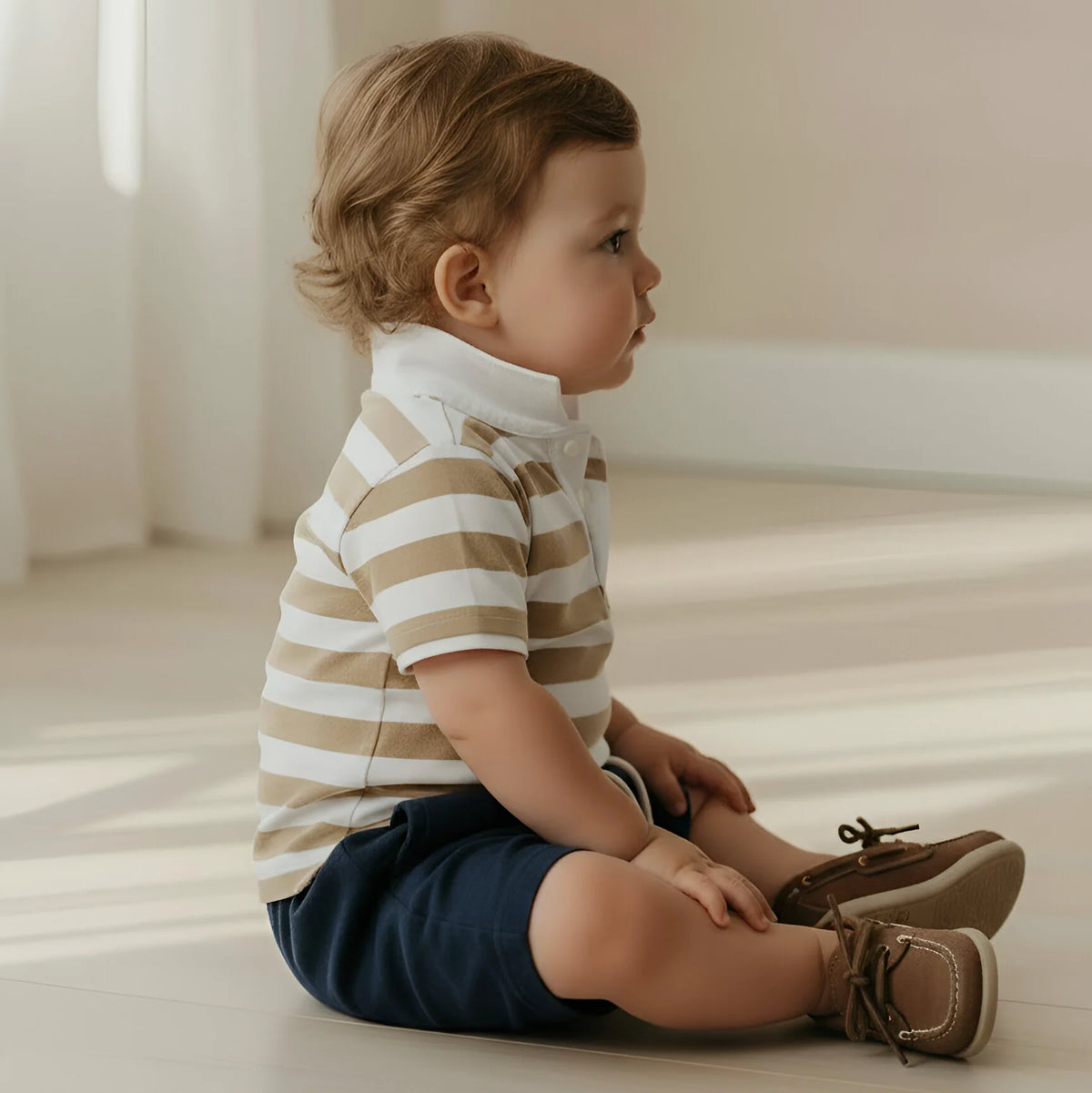 Child sitting on a light wooden floor wearing a striped shirt and shorts.