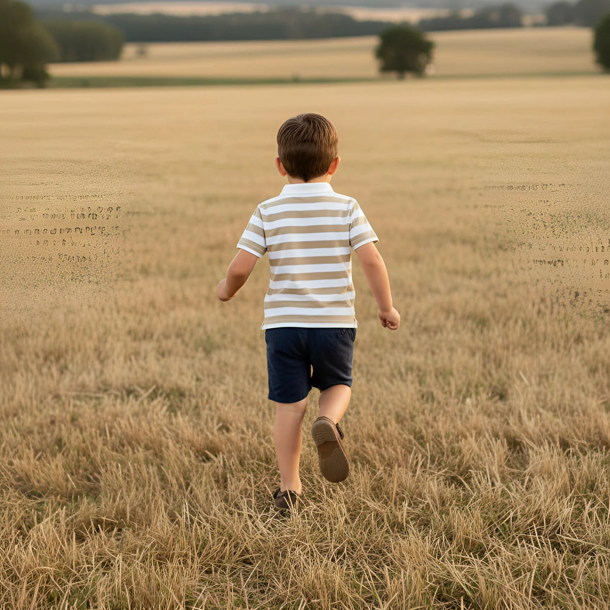 A young boy seen from behind walking through a dry grass field, wearing a cream and navy blue horizontally striped short-sleeve polo shirt and navy shorts.