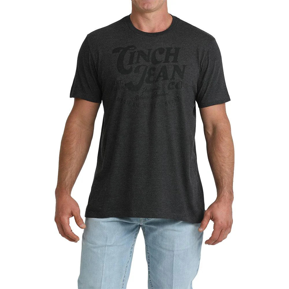 A man wearing a dark charcoal gray heather t-shirt with a faded black graphic print of 'Tinch Jean Co' on the chest, paired with light blue jeans, against a plain white background.