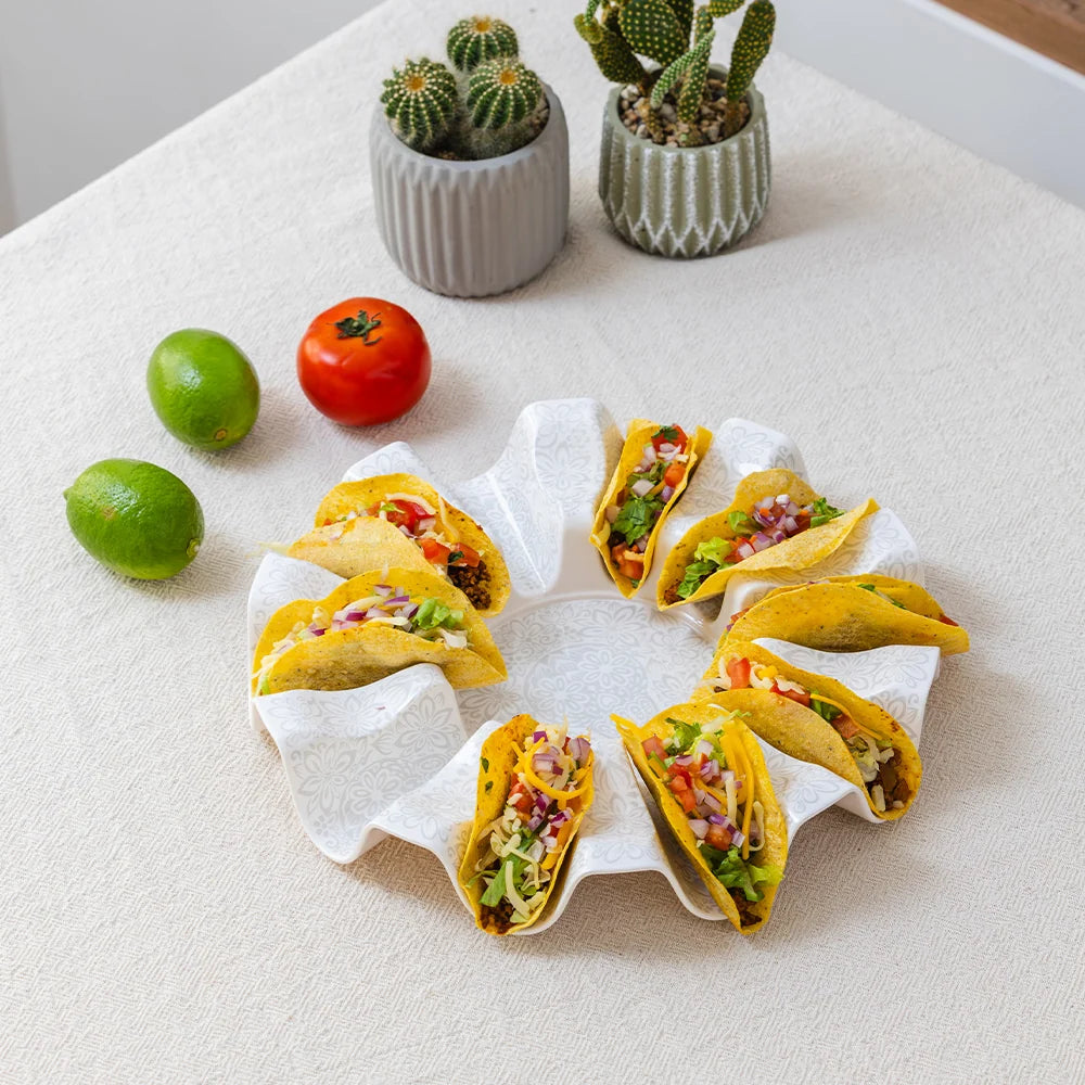 A ring-shaped white paper towel holder filled with yellow corn tacos, arranged on a light beige tablecloth with limes, a tomato, and two small potted cacti in the background.
