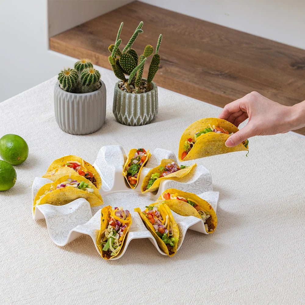 A hand holds a taco above a white wavy ring plate holding five tacos, with two small potted cacti and limes on a light tabletop.