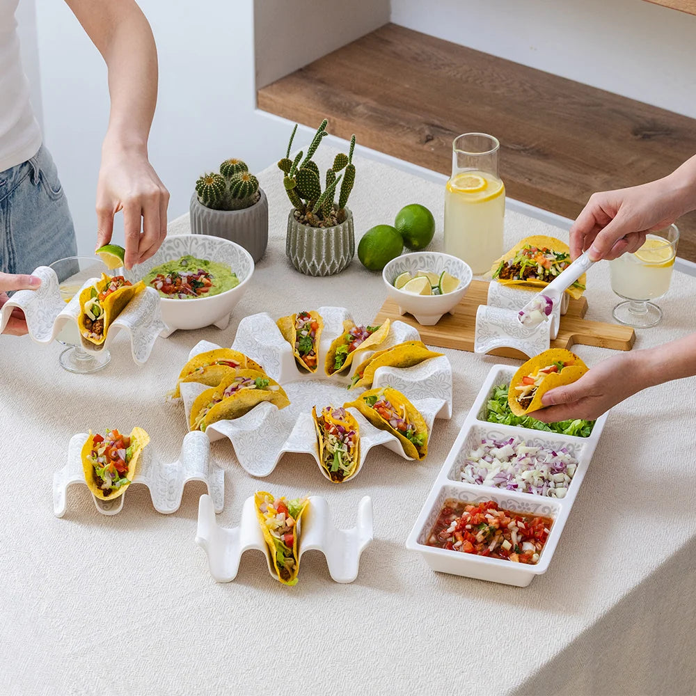White wavy ceramic taco holders arranged on a table, filled with tacos, alongside bowls of salsa, lime wedges, drinks, and small potted cacti. Multiple hands are visible serving food, indicating a lifestyle setting.