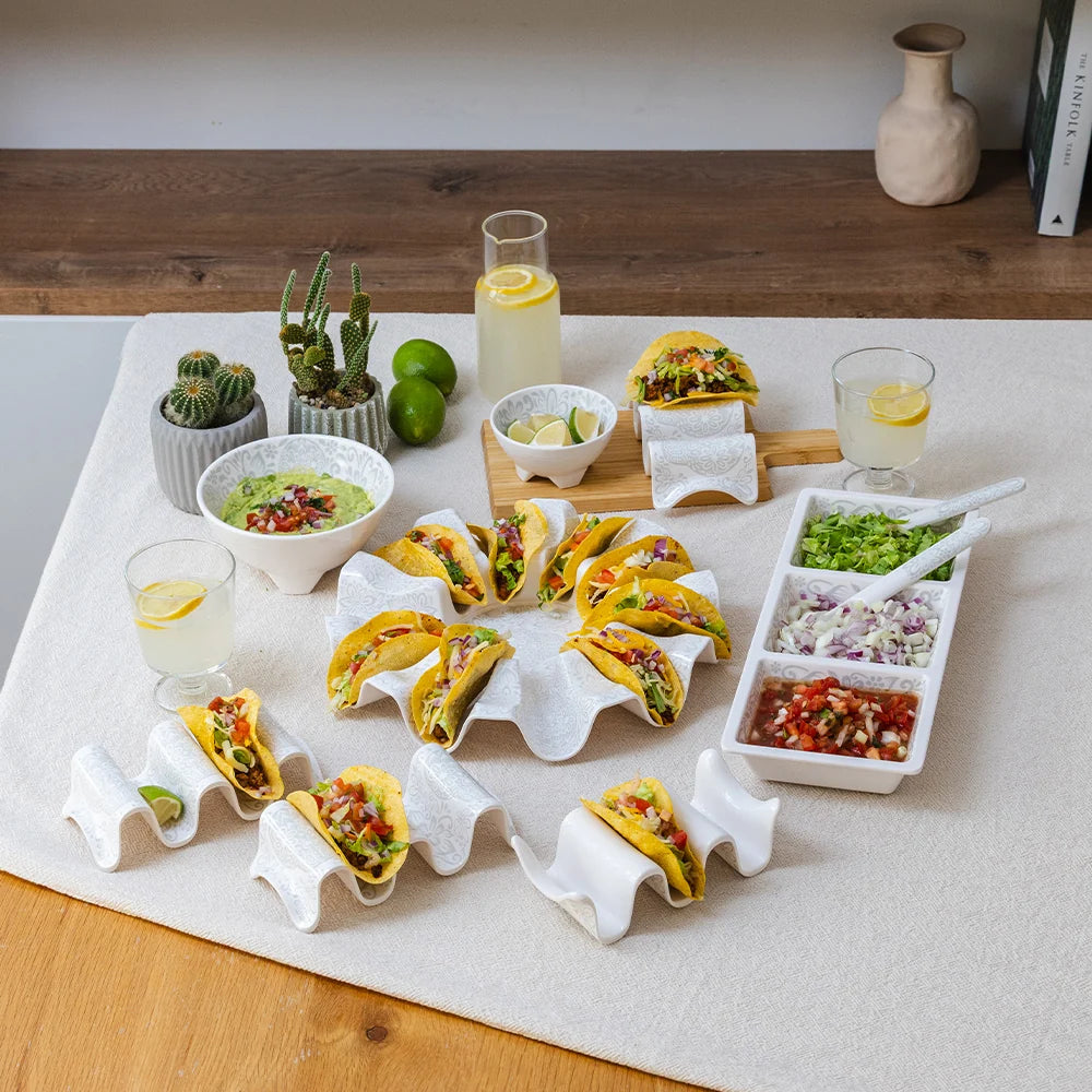 A styled food presentation on a white table runner, featuring multiple yellow hard-shell tacos arranged in wavy white paper holders, bowls of guacamole and salsa, limes, lemonade, and small potted cacti in the background.