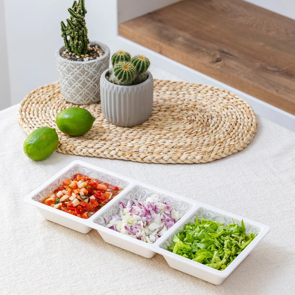 A three-compartment white plastic serving tray filled with chopped food, placed on a light fabric surface with a woven placemat, limes, and small potted cacti in the background.
