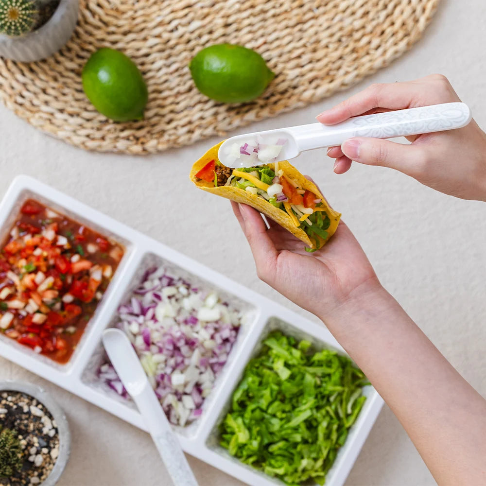 A person holds a taco while using a white plastic taco spoon to add salsa. In the foreground, a white divided tray holds chopped onions, shredded lettuce, and a red salsa, with two limes and a woven placemat in the background.