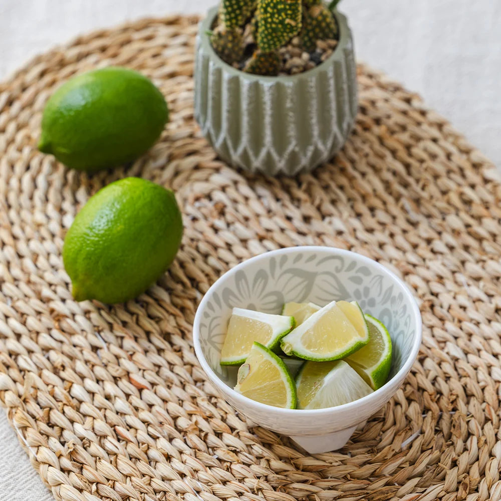 A small white ceramic bowl filled with lime wedges sits on a woven round placemat, with two whole limes and a small potted succulent in the background.