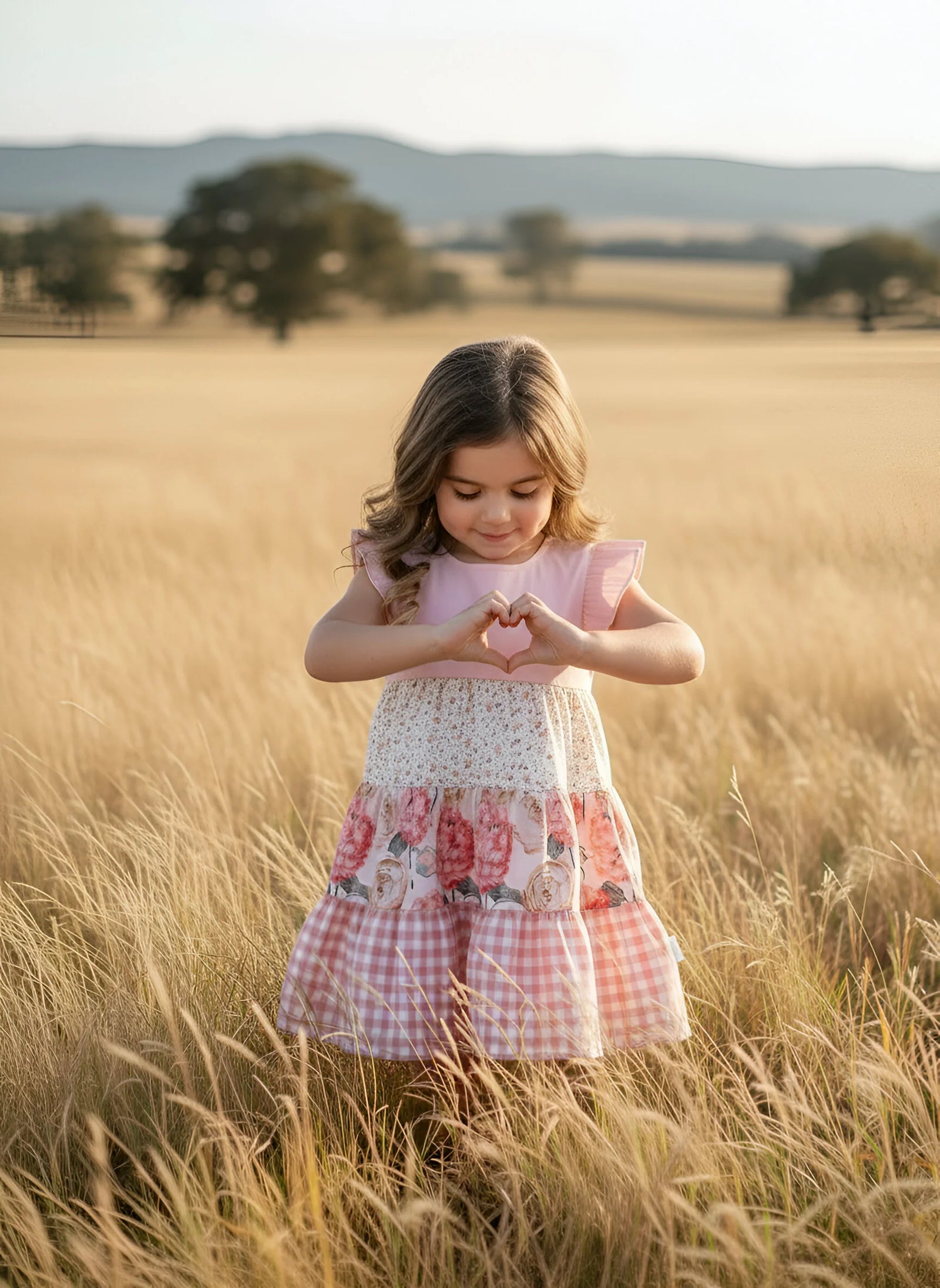 A young girl stands in a golden wheat field wearing a pink and white tiered dress with ruffle sleeves, a floral lace waist, and a pink gingham skirt.