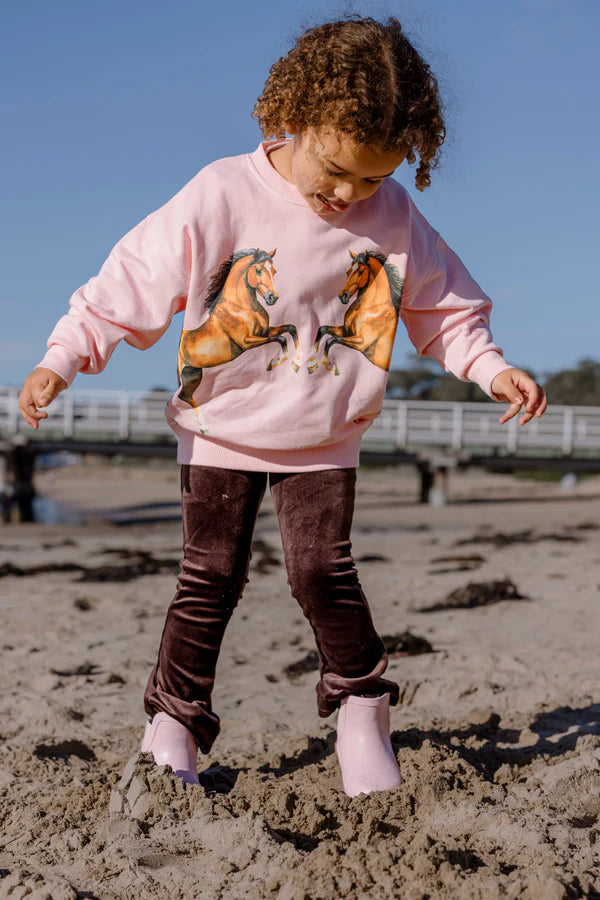 Child playing on a sandy beach wearing a pink sweatshirt with horse graphics and brown pants.