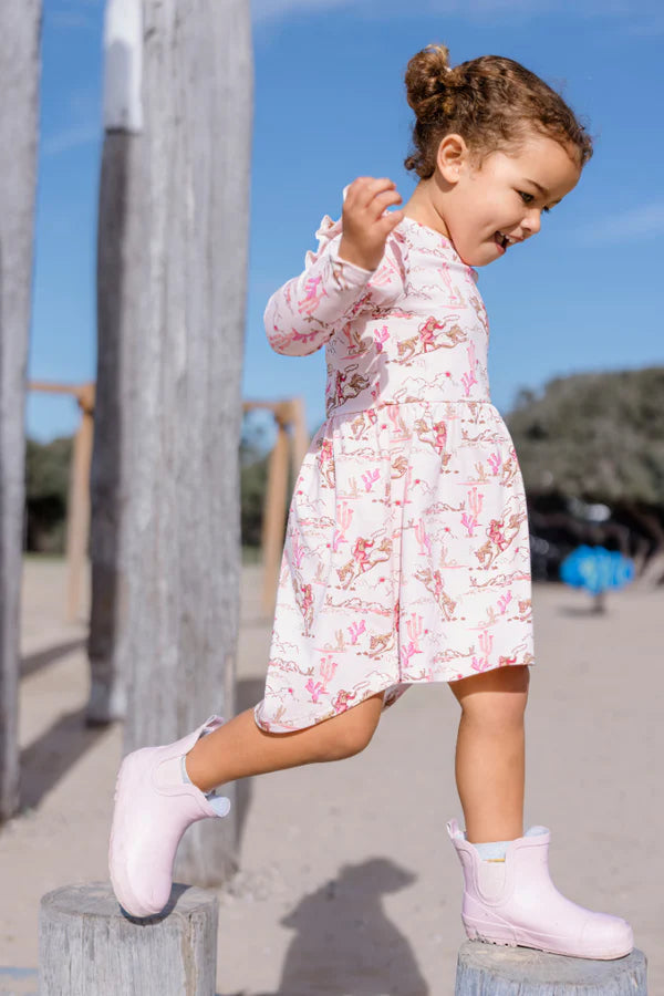 Child wearing a floral dress and pink boots standing on a concrete block with a blue sky background.