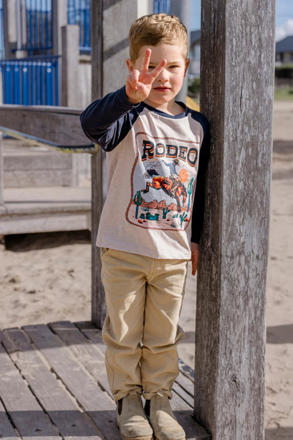 Child wearing a 'Rodeo' t-shirt standing near a wooden post outdoors.