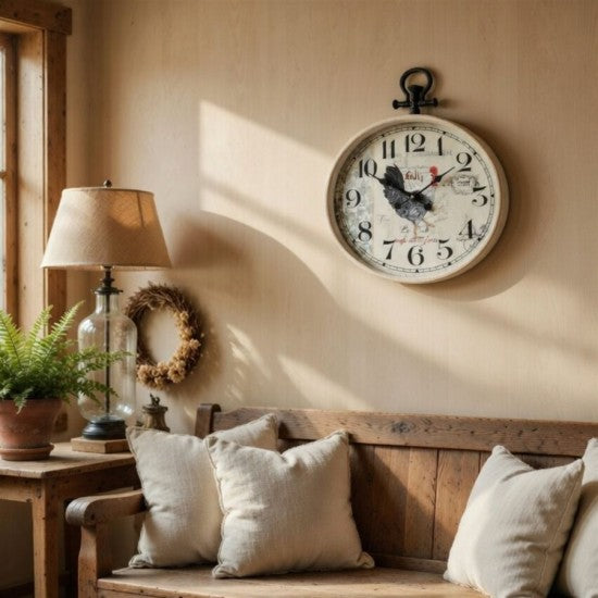 A round vintage-style wall clock with a distressed cream face, black numerals, and a rooster illustration, hanging on a beige wall above a rustic wooden bench with neutral throw pillows.
