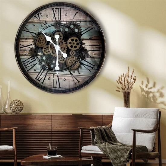 A large round wall clock with a rustic wood-grain face, black Roman numerals, and exposed gear details in the center, mounted on a beige wall above a wooden cabinet and white cushioned chair in a styled interior.