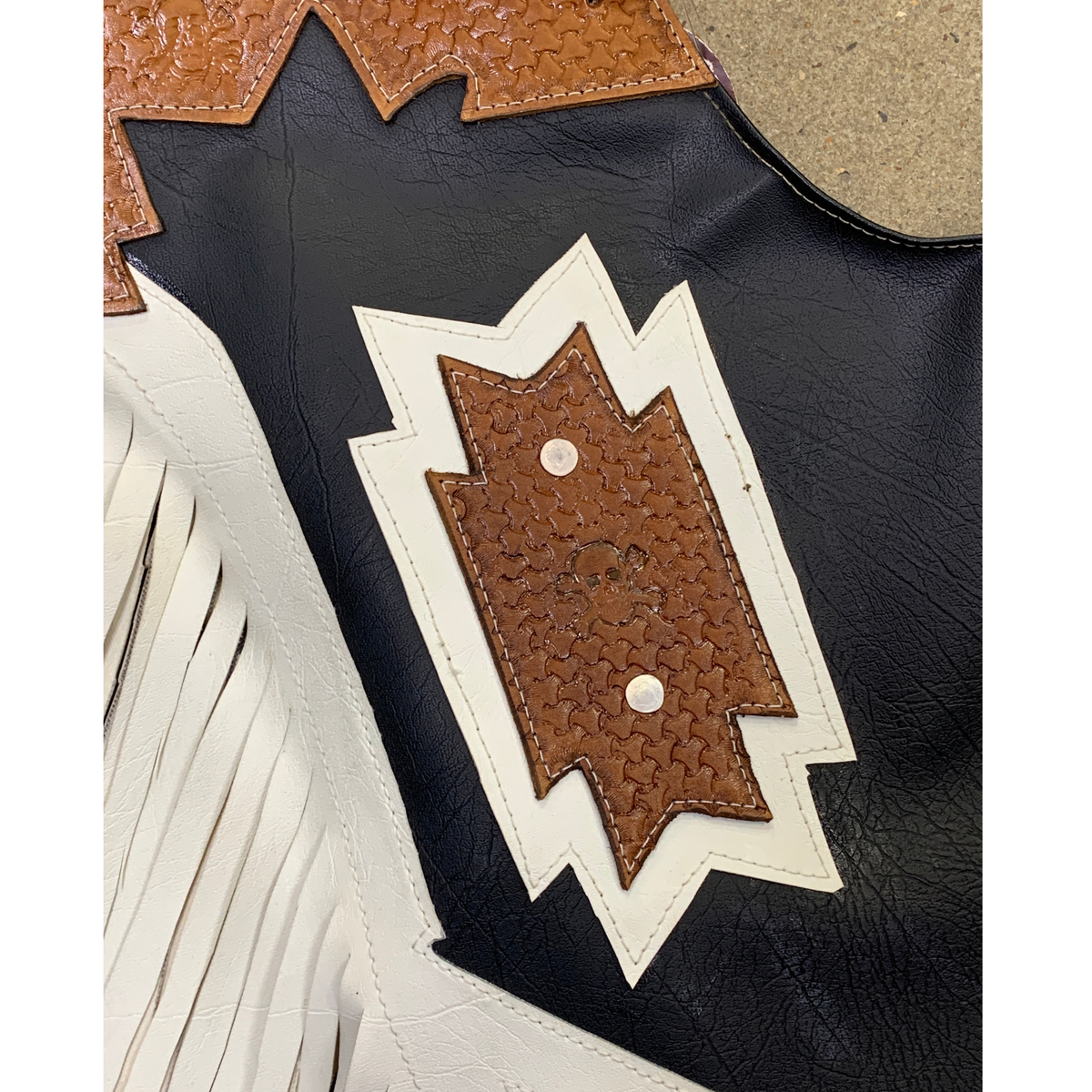 Close-up detail of a western-style leather boot or mule showing a black leather body, tan textured leather trim, white fringe, and a white-and-brown stitched decorative patch.