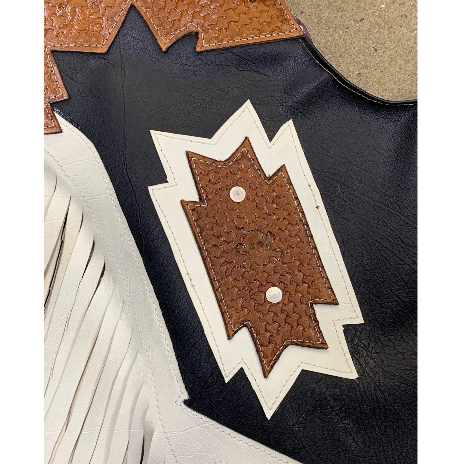 Close-up detail of a western-style leather boot or mule showing a black leather body, tan textured leather trim, white fringe, and a white-and-brown stitched decorative patch.