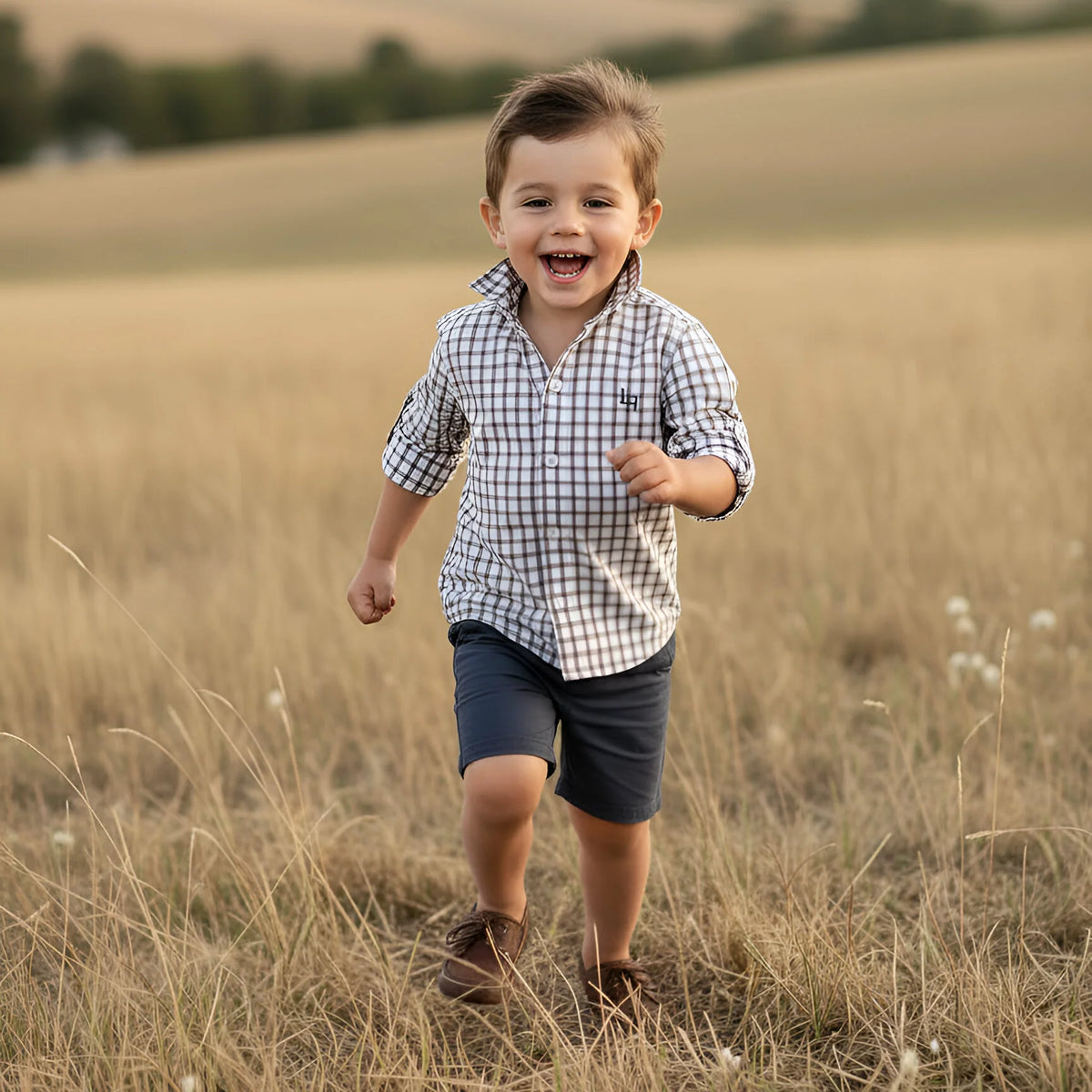 A young boy smiling and running through a dry grass field, wearing a white and navy blue gingham check long-sleeve shirt and dark navy shorts.