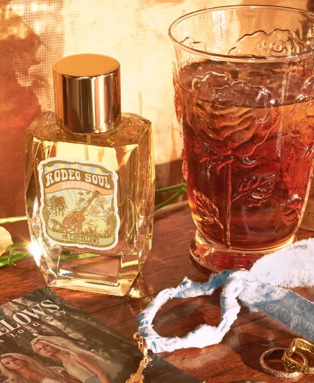 A bottle of Rodeo Soul cologne by True Scents next to a textured glass filled with an amber-colored drink on a wooden table, surrounded by decorative props including rings, a magazine, and a blue-and-white patterned cloth.