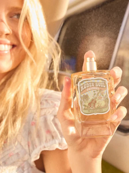A woman holds a bottle of Rodeo Soul Ranchero fragrance spray in her hand, with warm sunlight and a blurred car interior in the background.