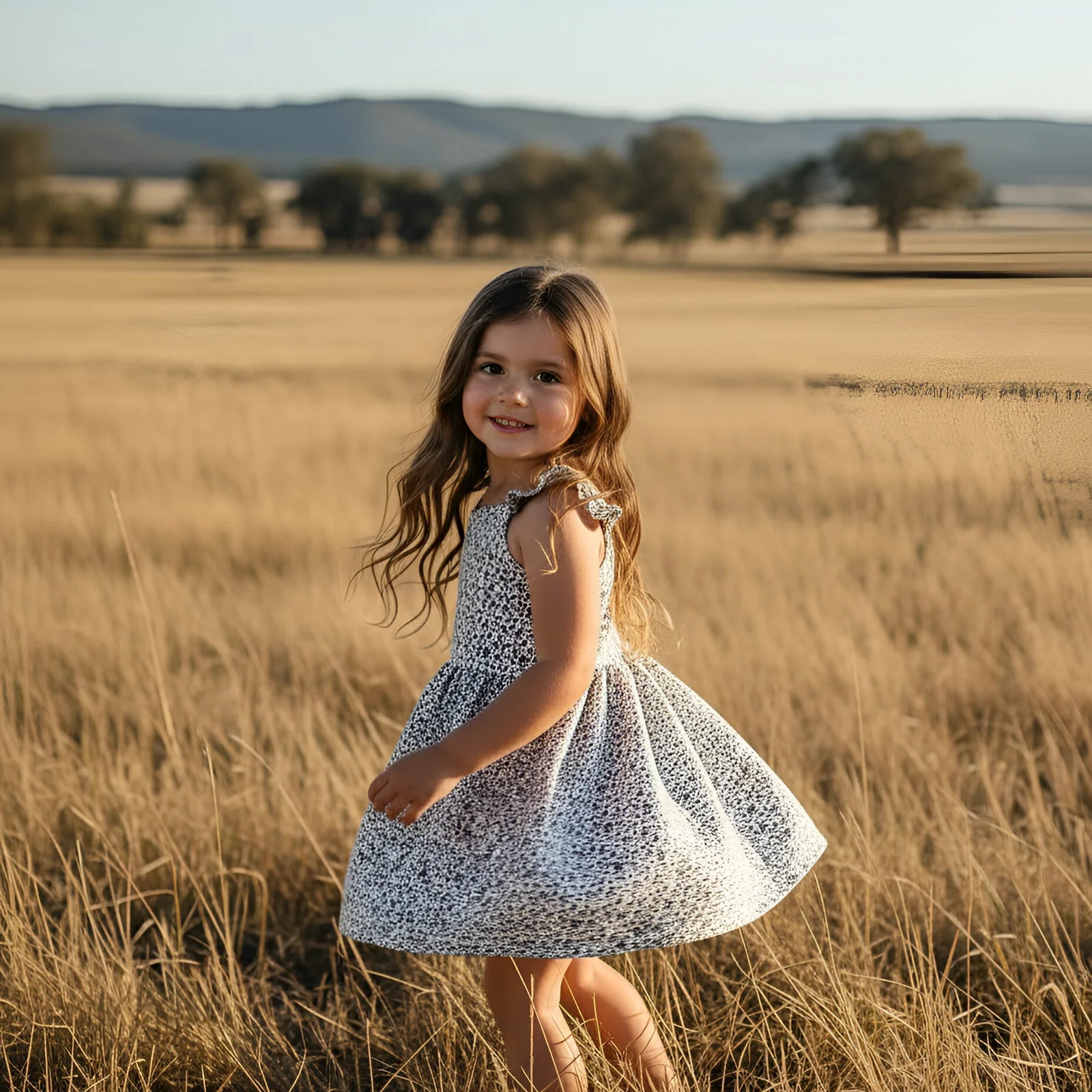 Young girl in a patterned dress standing in a field of tall grass with a scenic background.