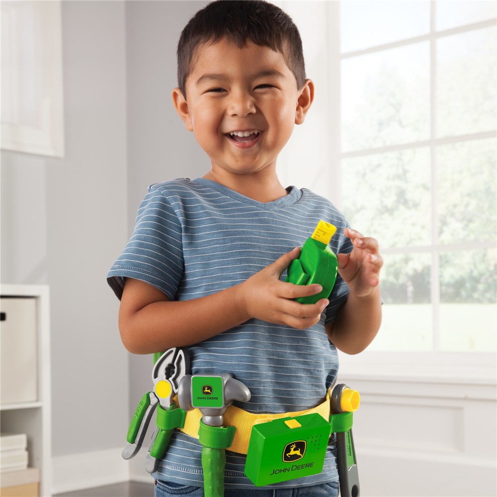 John Deere Deluxe Talking Toolbelt Set . Child wearing a toy tool belt with various tools, smiling indoors.