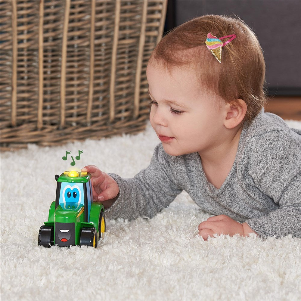A baby lying on a white shag rug playing with a green and blue toy tractor. The baby is wearing a gray long-sleeve shirt and has a small rainbow hair clip.