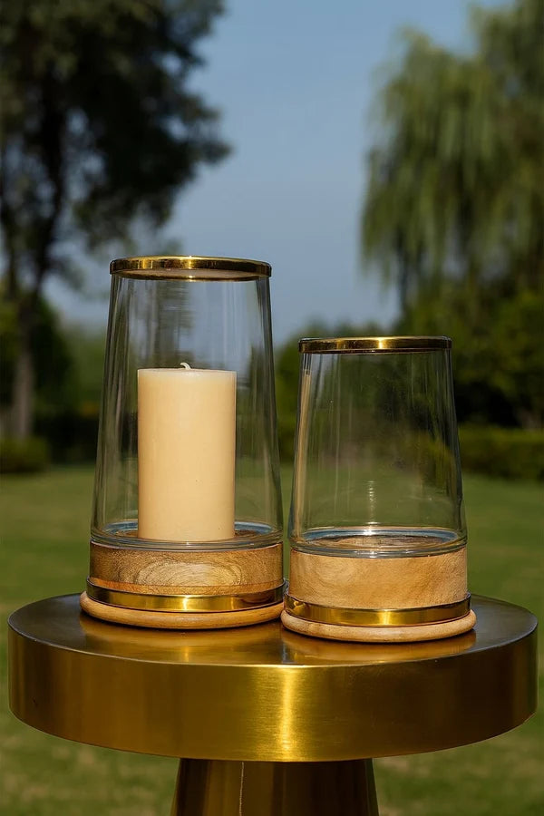 Two clear glass candle holders with gold trim and light wood bases, one containing a cream candle and the other empty, displayed on a round gold table outdoors with blurred greenery and blue sky in the background.