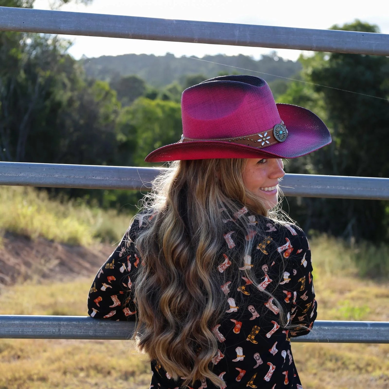 Person wearing a pink cowboy hat and patterned shirt standing behind a metal fence with trees in the background.