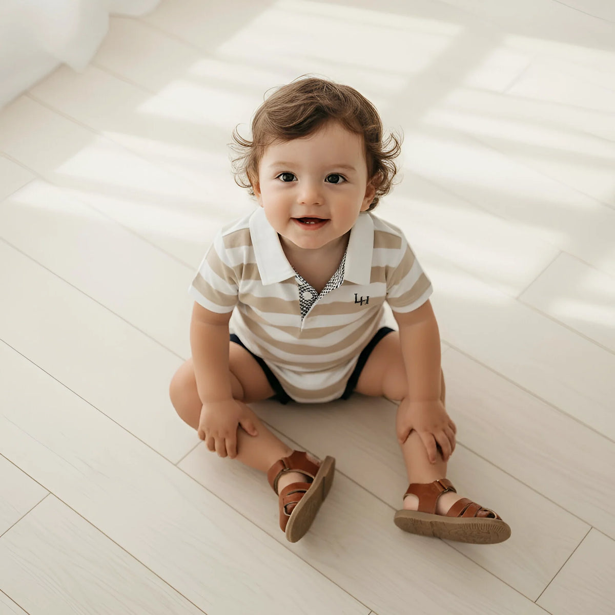 A toddler sitting on a light wood floor wearing a beige and white striped short-sleeve polo romper with brown sandals.