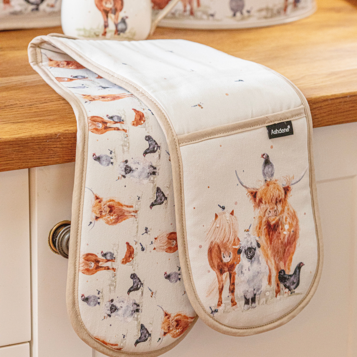 A kitchen oven mitt with a watercolor-style animal print featuring Highland cows, sheep, chickens, and birds, draped over a wooden countertop. The brand name 'Ashand' is visible on a small black label on the front pocket.