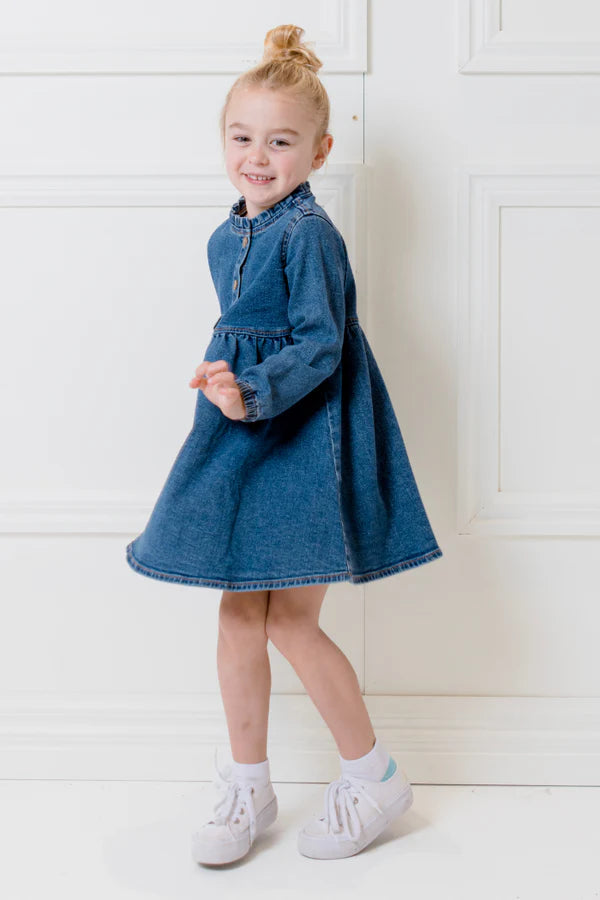 Young girl wearing a blue denim dress standing against a white paneled wall.