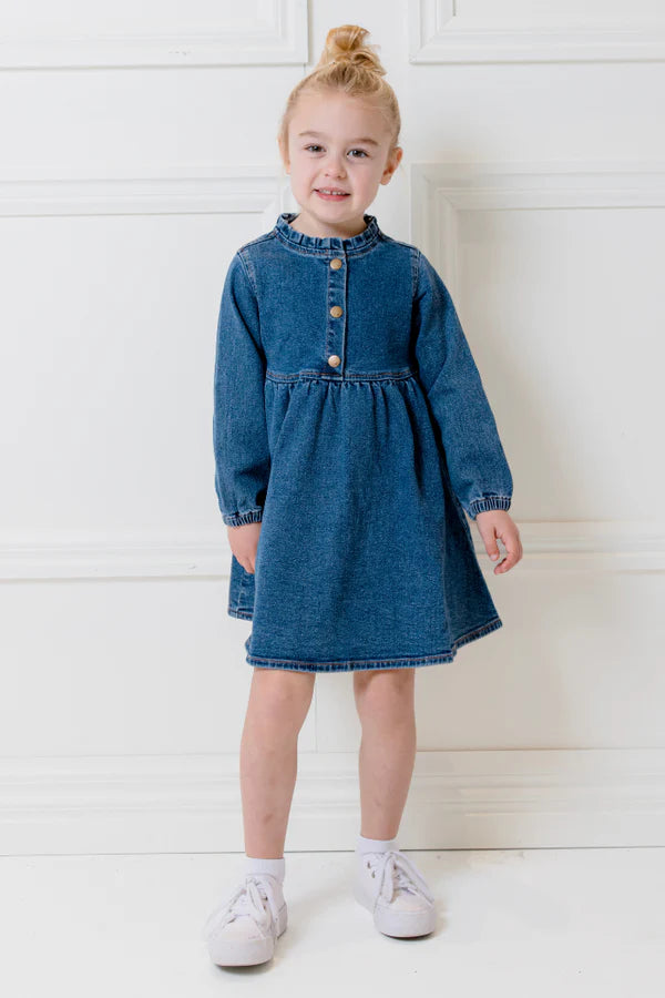 Child wearing a blue denim dress standing against a white wall.