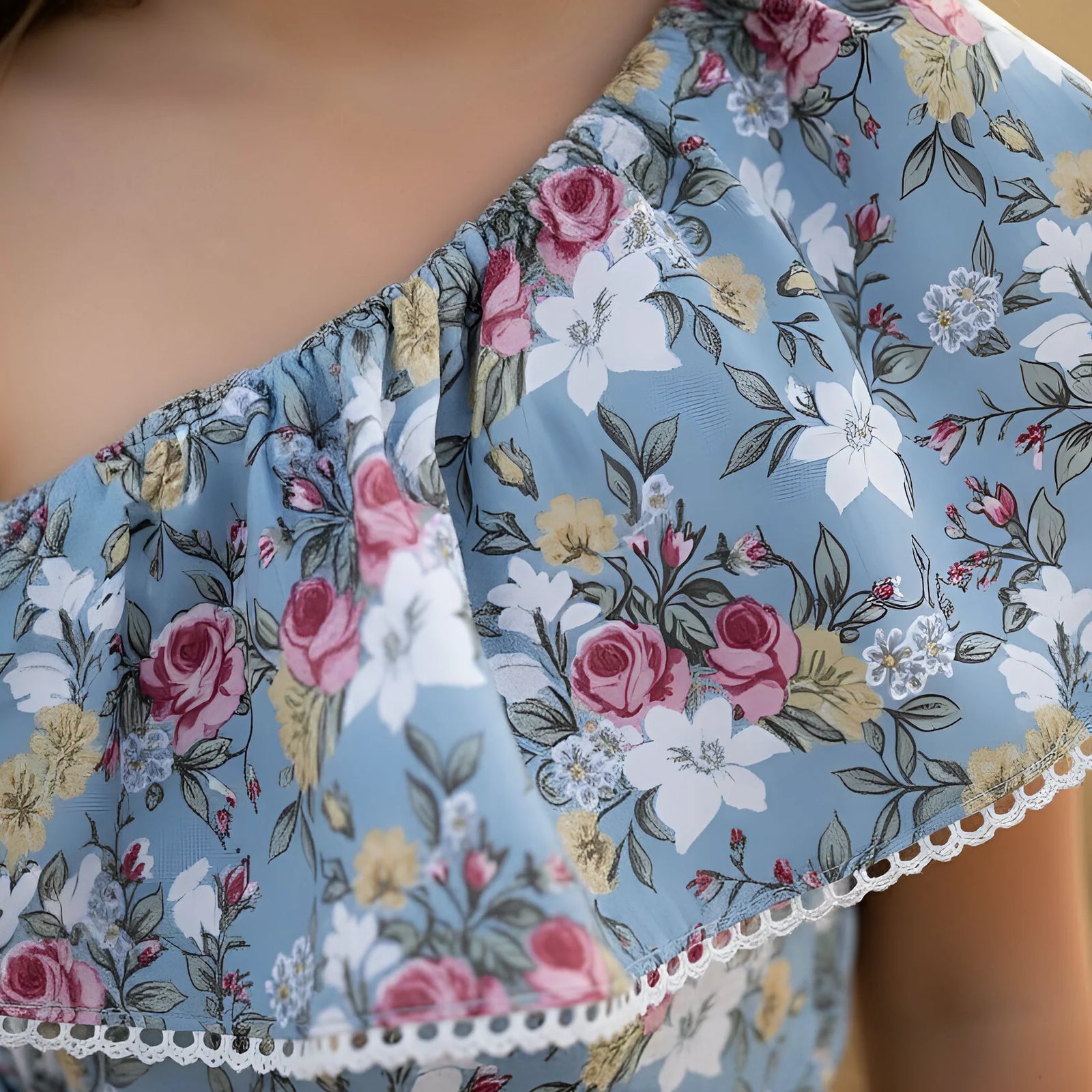 Close-up of a floral off-the-shoulder top with a light blue base, featuring pink, white, and yellow flowers with green leaves, finished with a white lace trim.