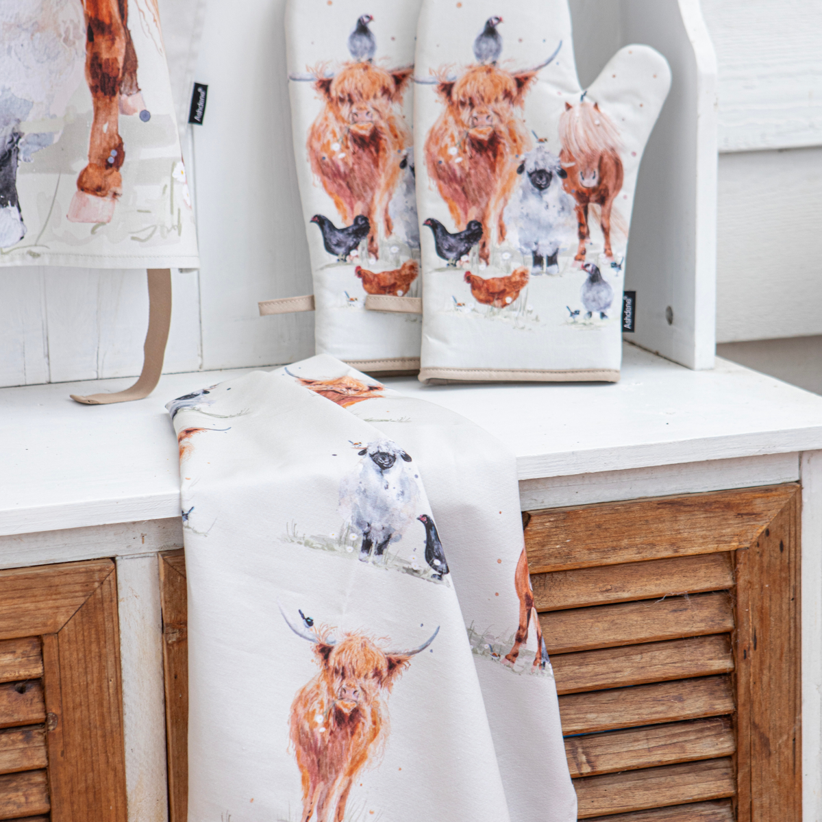 A white kitchen textile set featuring a watercolor-style print of Highland cows, sheep, chickens, and birds, displayed on a white shelf with wooden louvered cabinets behind it. The set includes a towel or cloth draped over the front and matching oven mitts and a hanging cloth behind.