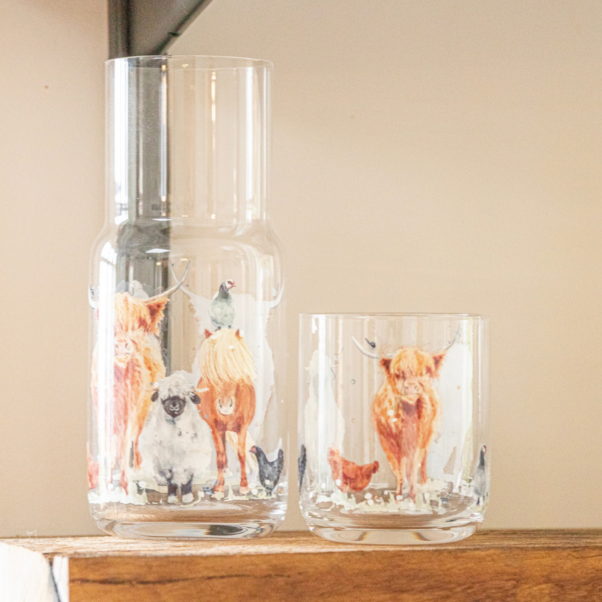 A clear glass carafe and matching lowball glass decorated with painted Highland cows, sheep, chickens, and birds, displayed on a wooden surface against a beige wall.