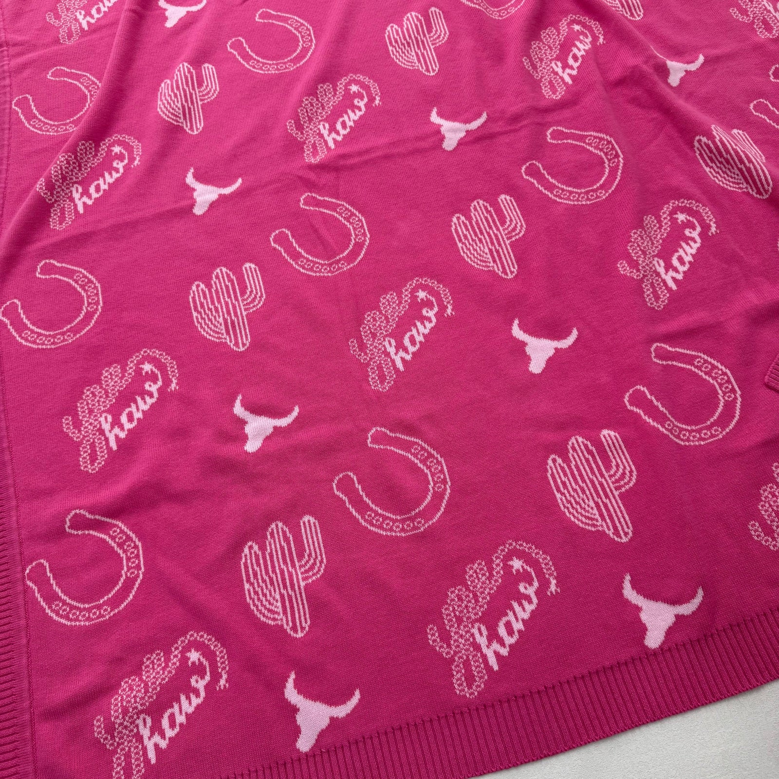 A close-up of a bright pink knit fabric with a repeating white western-themed print featuring cacti, horseshoes, longhorn silhouettes, and the word 'Haws' in script.