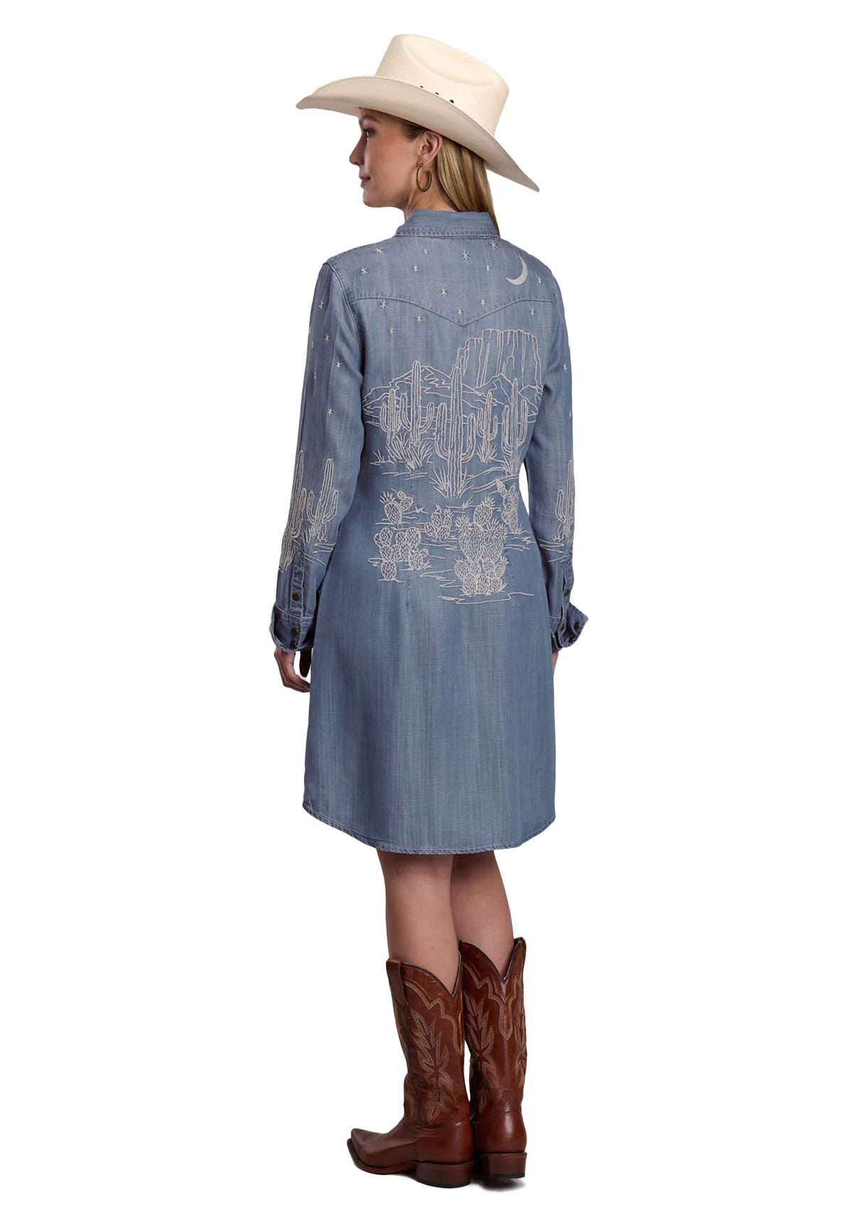 Woman wearing a blue denim dress with white embroidery, brown cowboy boots, and a white cowboy hat on a white background.