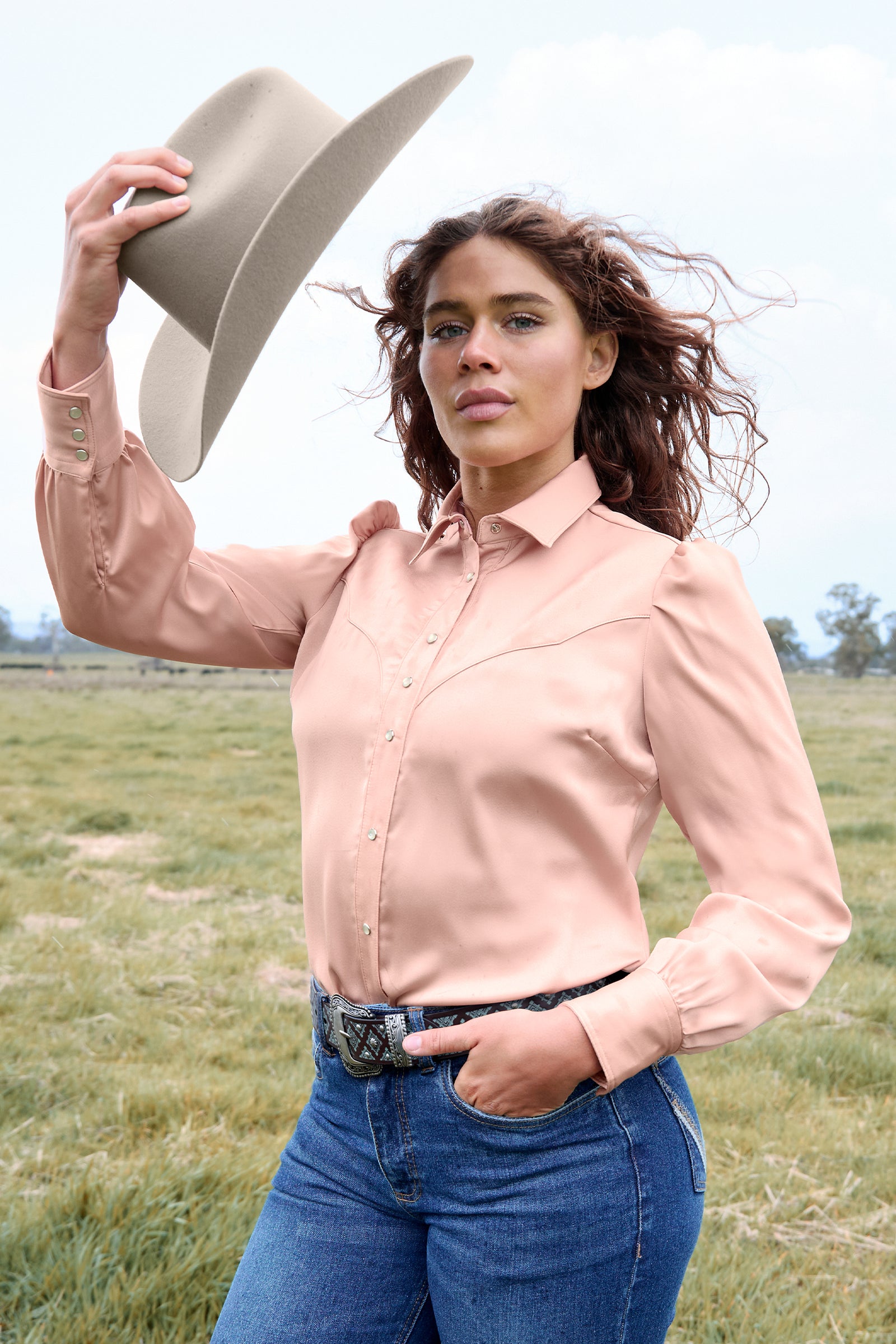 Woman in a pink shirt and blue jeans holding a beige cowboy hat in an open field.