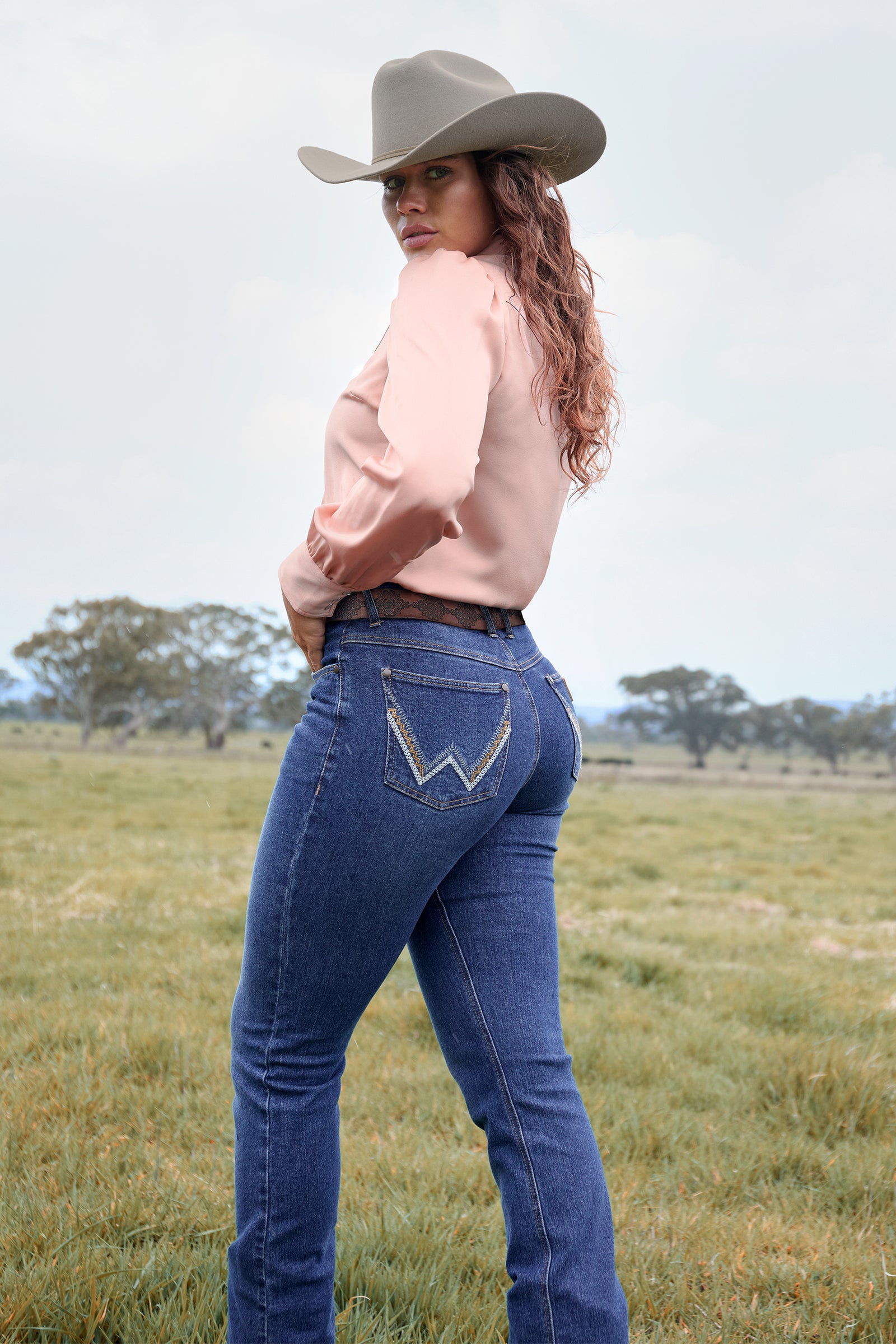 Woman wearing blue jeans and a beige cowboy hat in an open field.