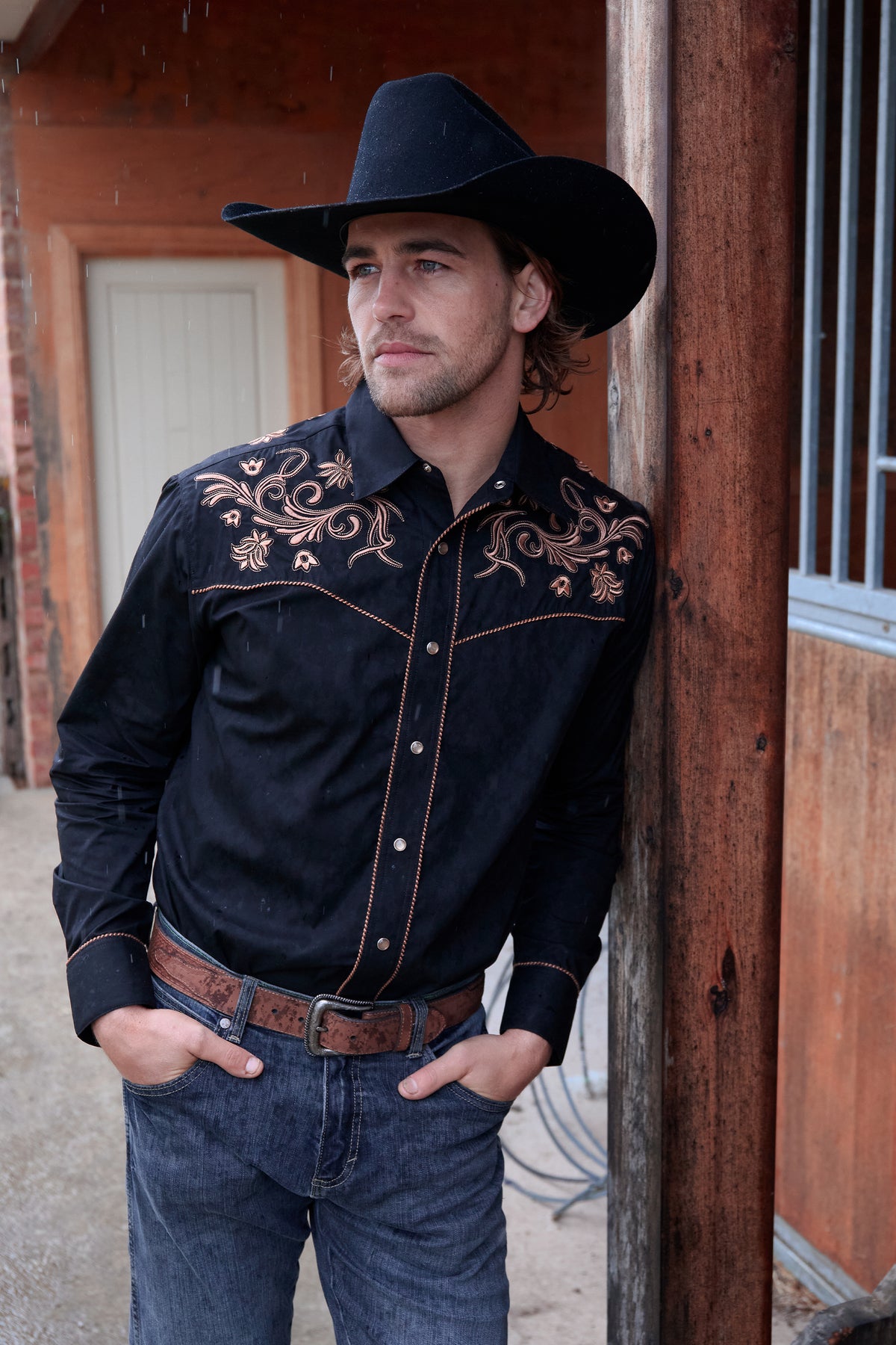 Man wearing a black embroidered shirt and cowboy hat leaning against a wooden building.