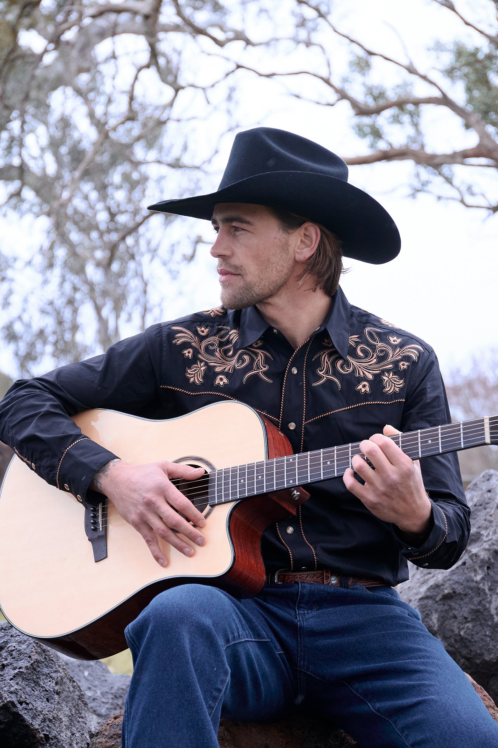 Man in cowboy hat playing guitar outdoors with trees in the background