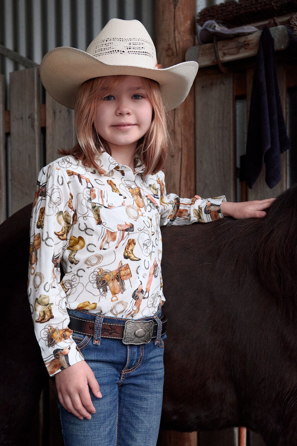Young girl in a cowboy hat and patterned shirt standing next to a horse in a barn setting.
