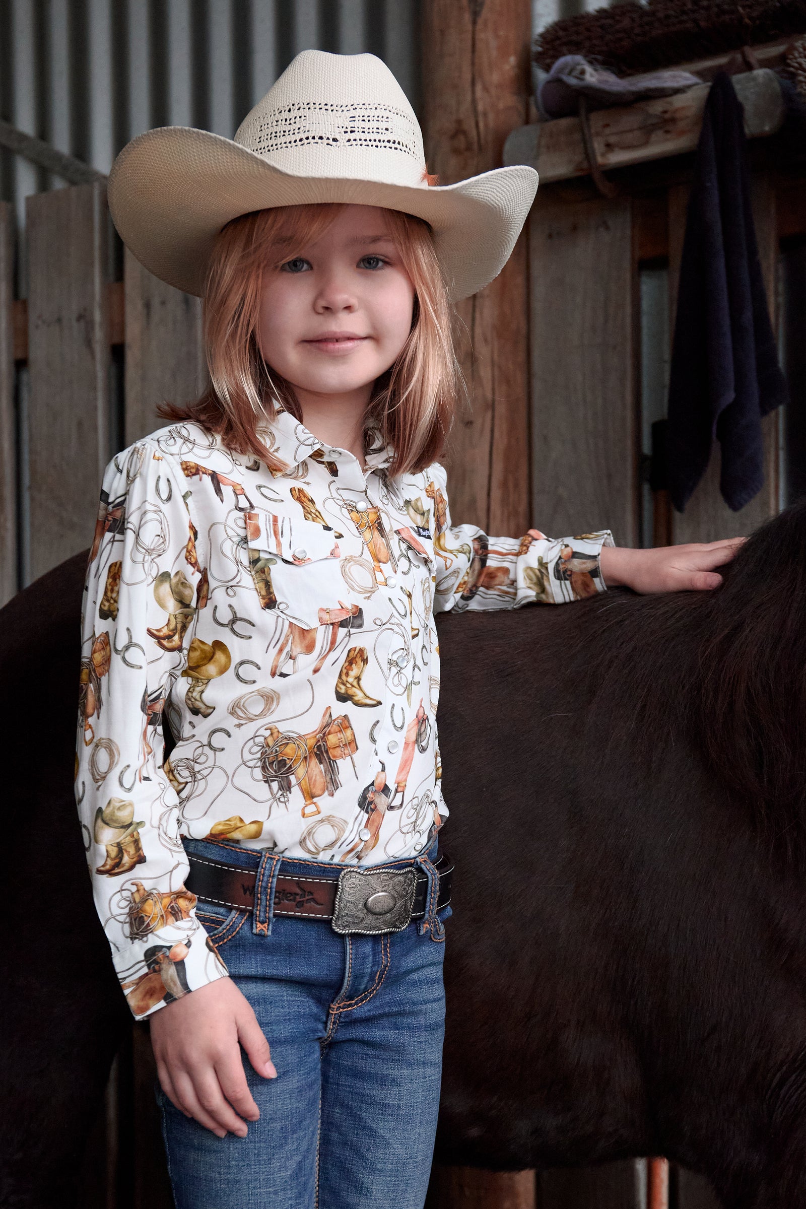 Young girl in a cowboy hat and patterned shirt standing next to a horse in a barn setting.