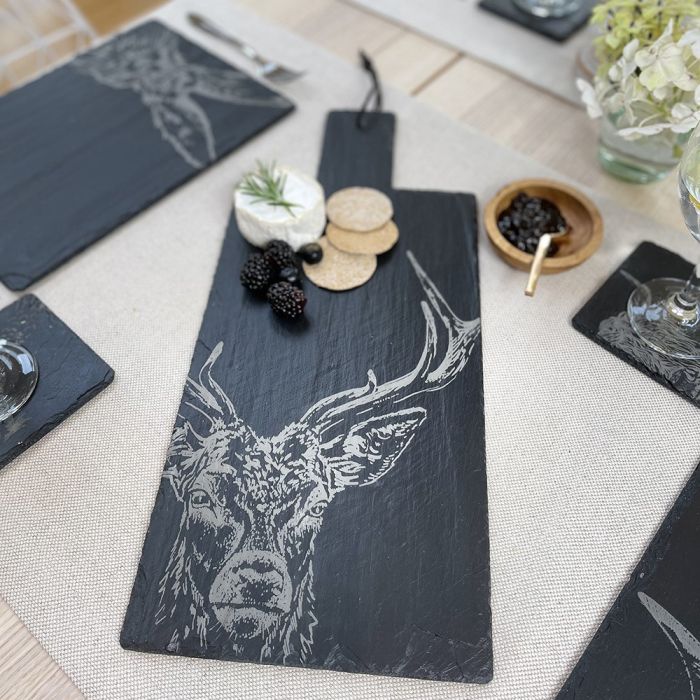 A rectangular black slate serving board with a white engraved deer head design, styled on a beige tablecloth with cheese, crackers, berries, and other place settings.