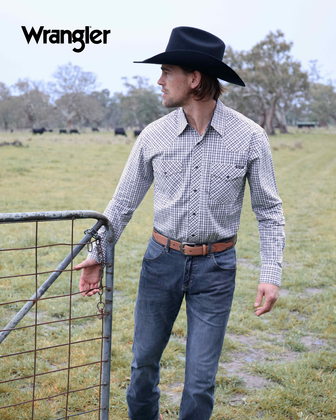 Man wearing a checkered shirt and jeans standing next to a gate with Wrangler branding.