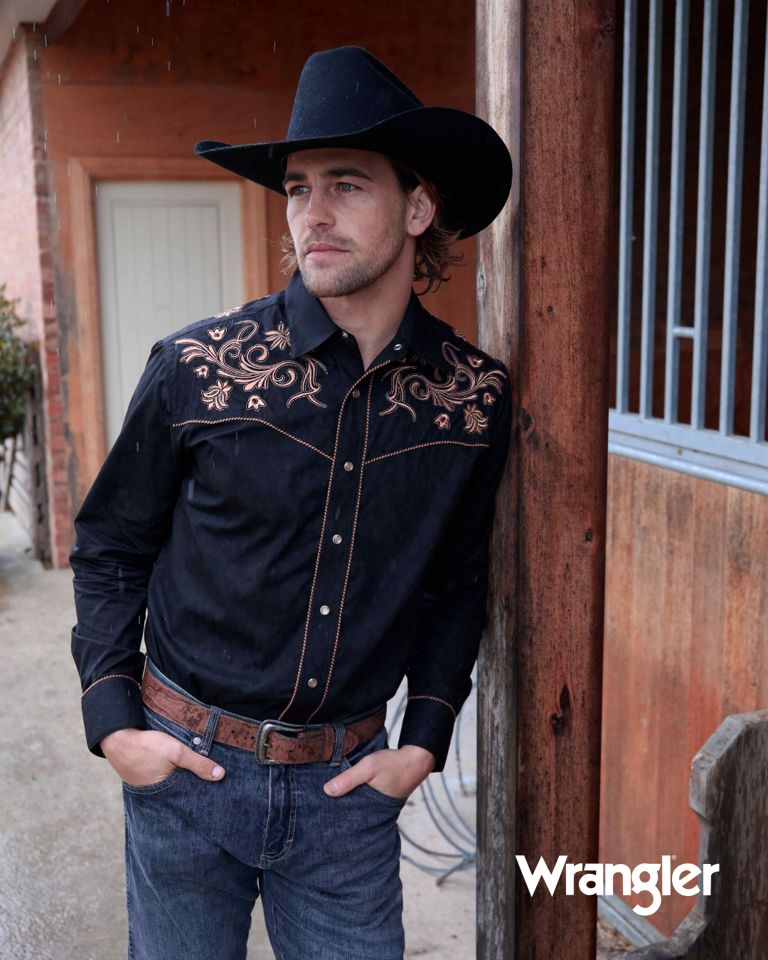 Man wearing a black embroidered shirt and cowboy hat leaning against a wooden building, with 'Wrangler' branding.