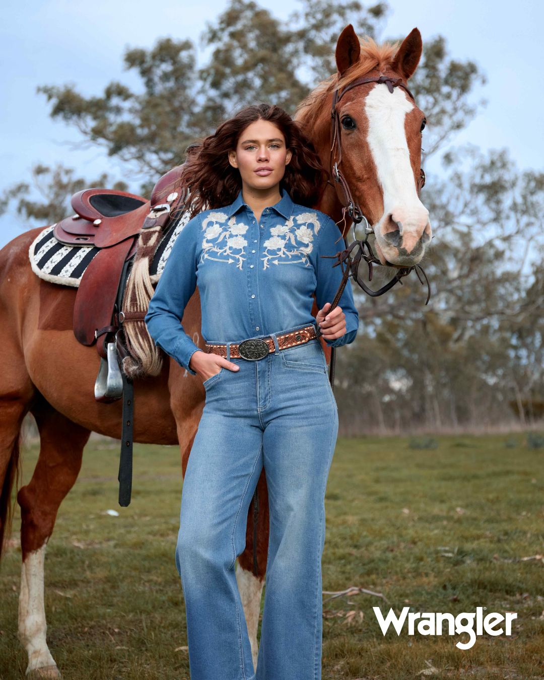 Woman in a blue jumpsuit standing next to a horse with Wrangler branding.