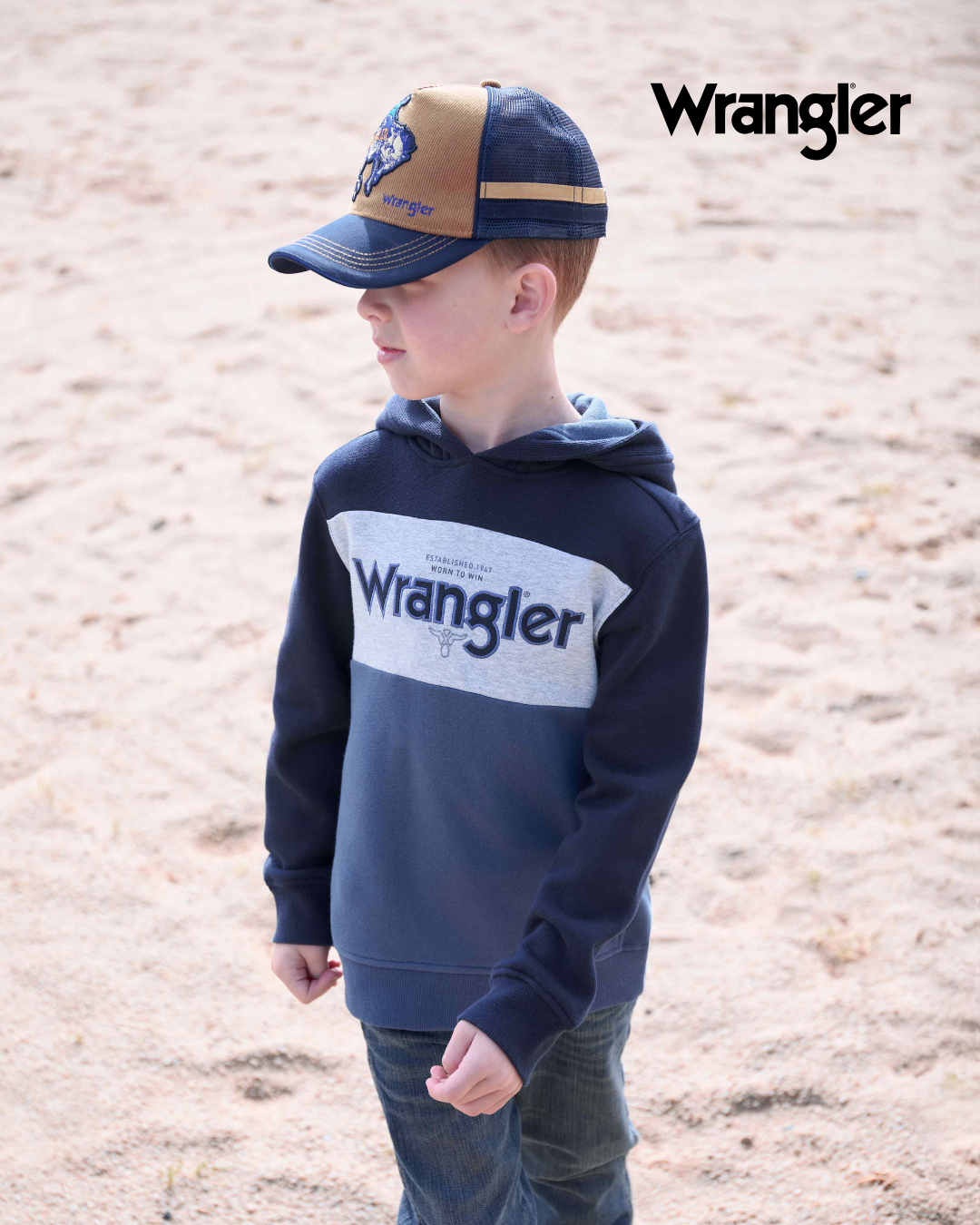Child wearing a navy hoodie with Wrangler logo and cap on a sandy background