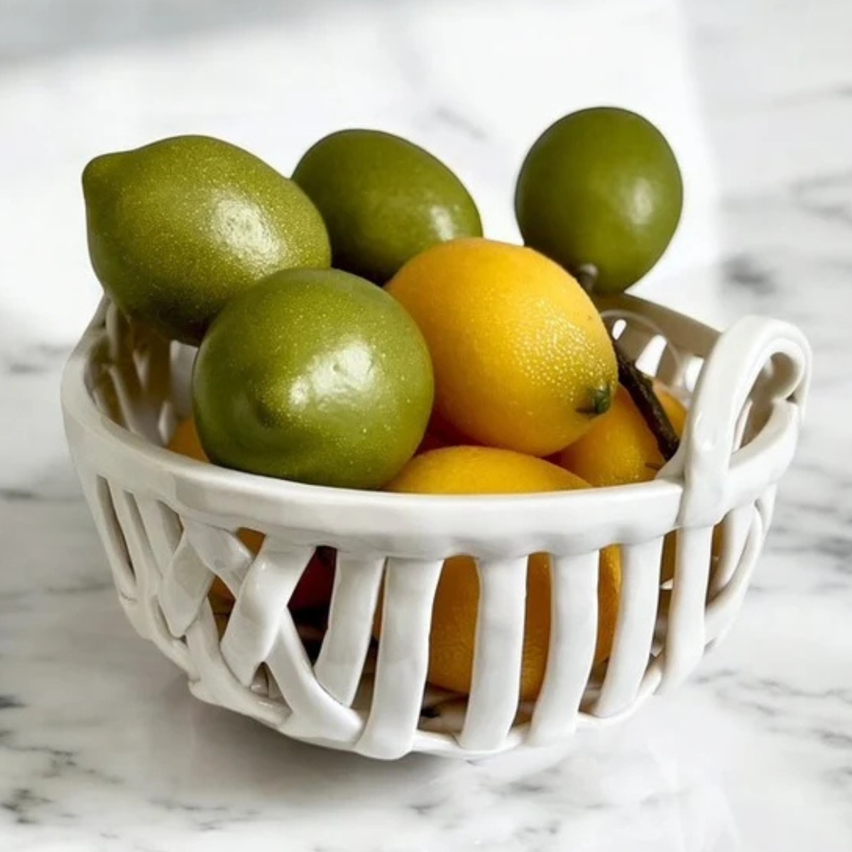 A white ceramic fruit basket filled with green and yellow lemons, set on a white marble surface.